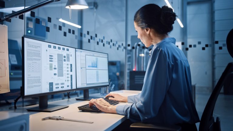 Ingenieurin arbeitet an zwei Monitoren mit technischen Plänen in einer Industriehalle